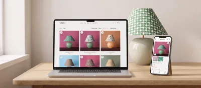 Salig Studios webbshop på en laptop och en mobil, bredvid en av deras bordslampor